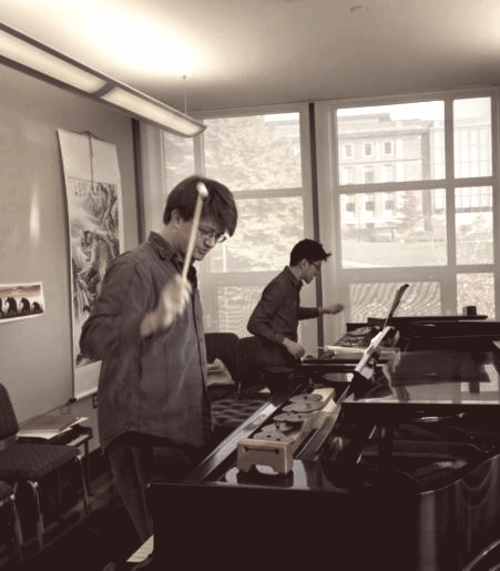 Duo rehearsing a two-piano work; both are raising mallets about to strike two woodblocks