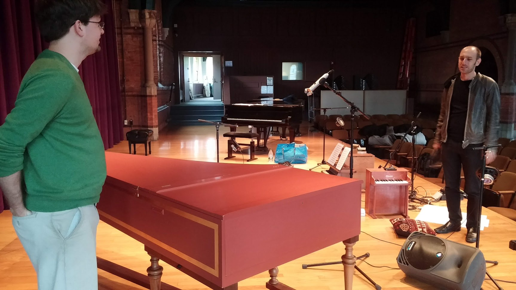 Photo of two people on a stage full of gear: a harpischord in front, speakers, mic stands, a toy piano, and a grand piano in the back.